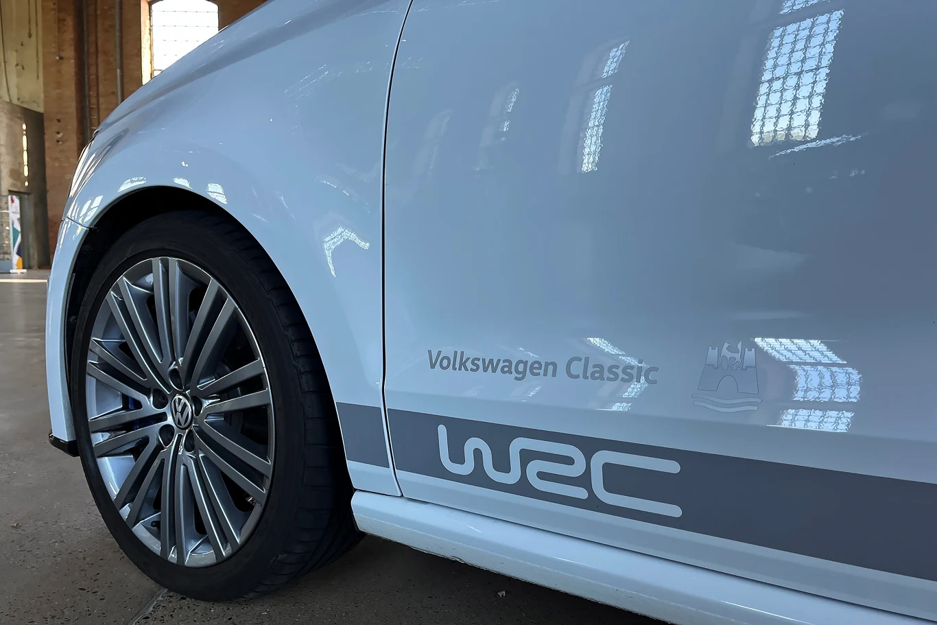 Volkswagen Polo R WRC wyraźnie zaznaczał swoją przynależność do rajdowej rodzinny Volkswagena. Biały samochód Volkswagen Classic z widocznym logo WRC na bocznej części nadwozia. Auto stoi wewnątrz budynku z cegły, przez okno wpada światło, które odbija się na lakierze samochodu. Stylowe felgi dodają sportowego wyglądu.