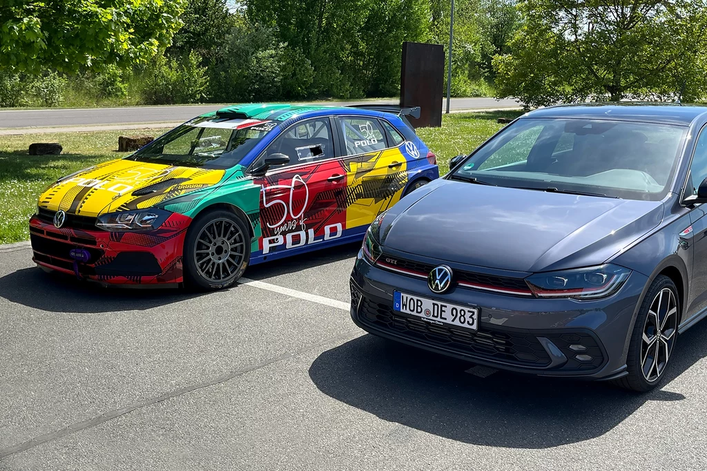 Rajdowe Polo R5 oraz jego cywilny odpowiednik po faceliftingu. Podobne, jednak w rzeczywistości zupełnie różne pojazdy. Dwa samochody Volkswagen zaparkowane obok siebie na parkingu. Jeden ma wielobarwne malowanie i napisy, drugi jest tradycyjnie szary. W tle widoczne są drzewa i fragment drogi.
