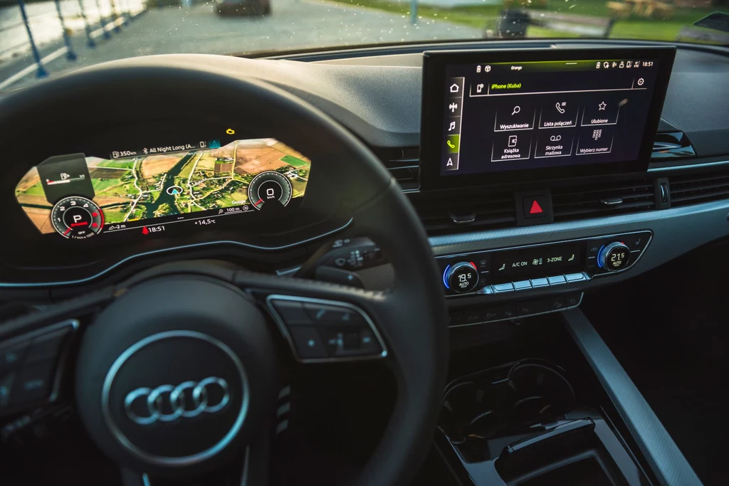 Audi A4 B9 może pochwalić się zwykle bogatym wyposażeniem, w tym tak zwanym wirtualnym kokpitem z możliwością personalizacji wyglądu zegarów Wnętrze samochodu marki Audi z widocznym zaawansowanym panelem sterowania, wyświetlaczem nawigacji oraz ekranem dotykowym. Kierownica z logotypem producenta, a na tapicerce widoczna wysokiej jakości estetyka wykończenia.