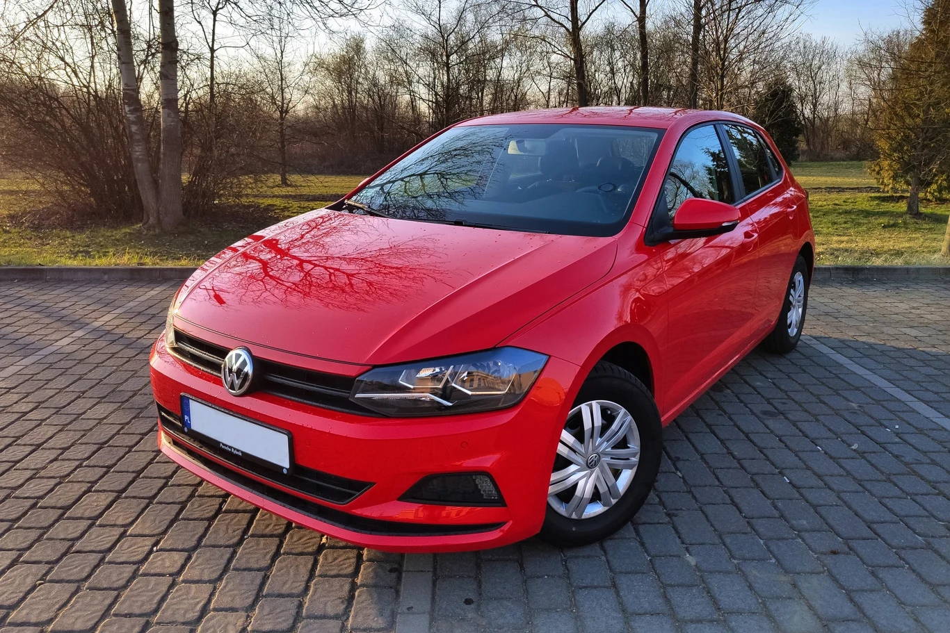 Polo VI urosło, czyniąc z niego samochód podobny rozmiarami do Volkswagena Golfa IV generacji. Czerwony samochód marki Volkswagen Polo zaparkowany na kostce brukowej w otoczeniu drzew w tle.