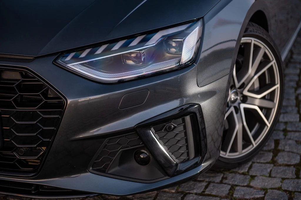 Szukając używanego Audi A4 B9 warto postawić na dwa elementy wyposażenia - pakiet S line oraz matrycowe reflektory Przedni reflektor nowoczesnego samochodu z charakterystycznym wzorem świateł, widoczna elegancka karoseria i alufelgi na tle brukowanej nawierzchni.