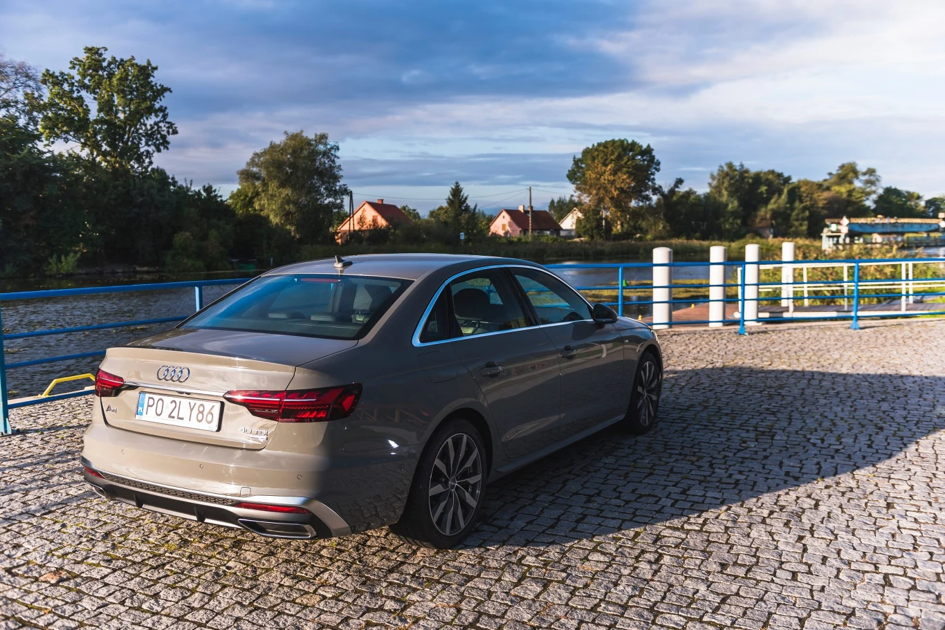 Samochód Audi stoi na brukowanym parkingu, w słoneczny dzień. W tle widać zieloną roślinność, domy oraz zbiornik wodny, co tworzy spokojny, malowniczy krajobraz.