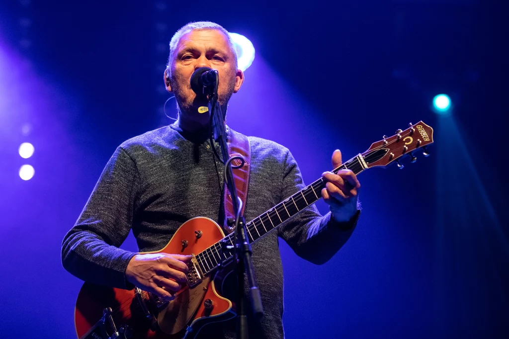 Mężczyzna grający na gitarze na scenie, w otoczeniu efektów świetlnych. Jego wyraz twarzy jest skupiony, a otoczenie tworzy atmosferę koncertu na żywo.