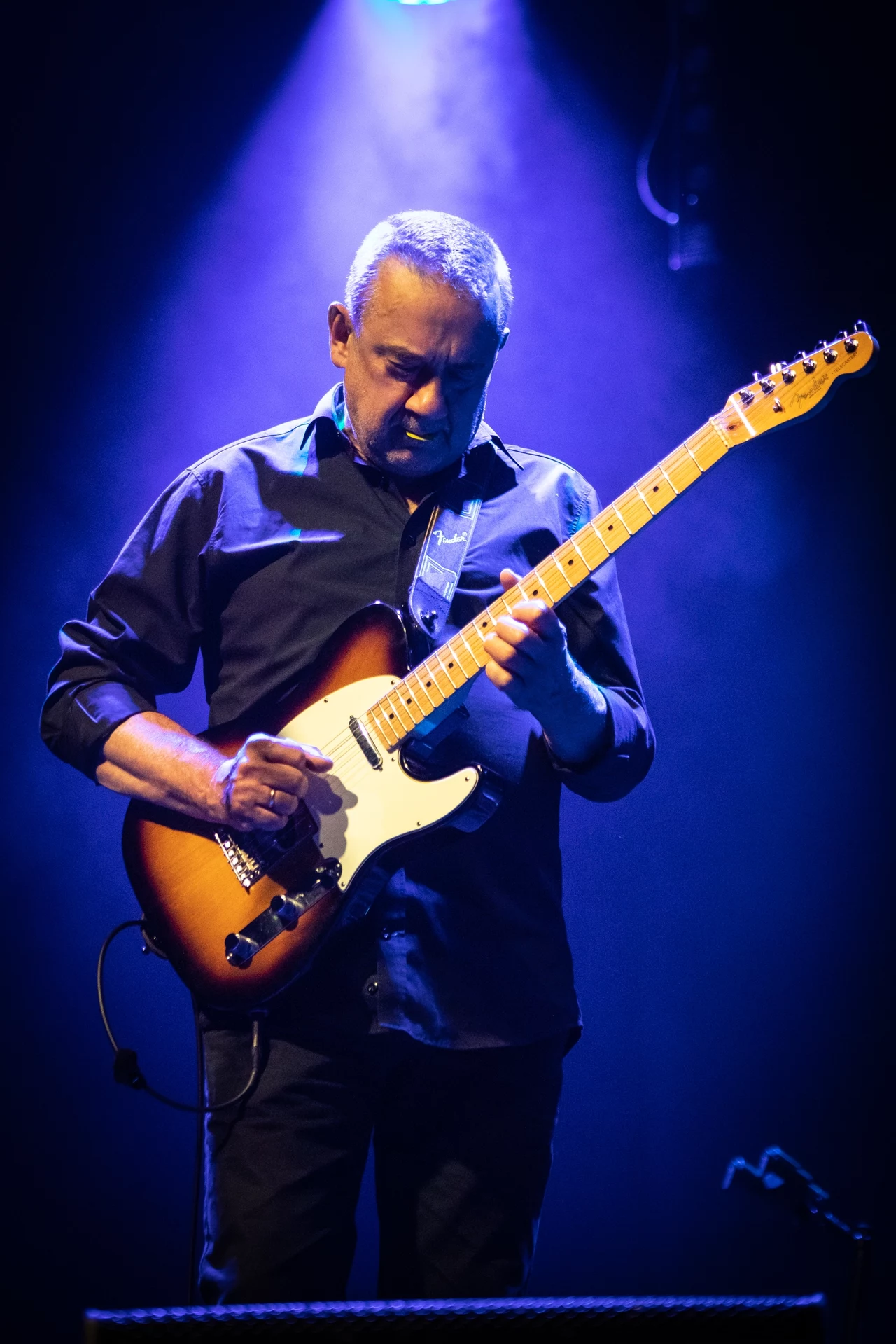 Mężczyzna grający na gitarze elektrycznej na scenie, oświetlony niebieskim światłem.