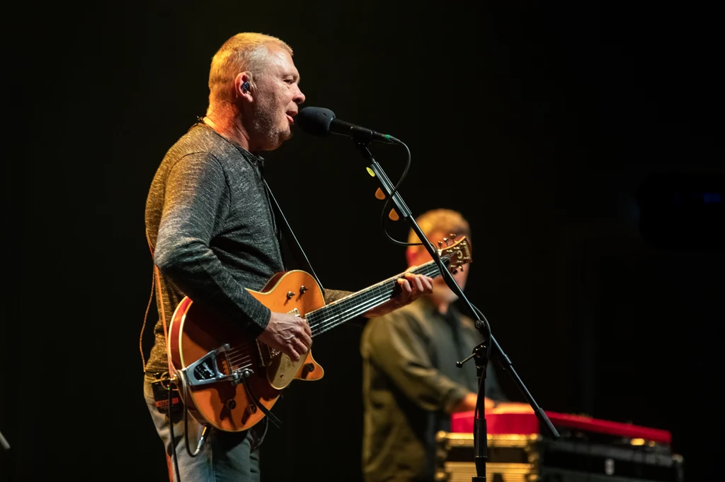 Mężczyzna śpiewający i grający na gitarze elektrycznej na scenie podczas koncertu, w tle drugi mężczyzna przy klawiaturze.