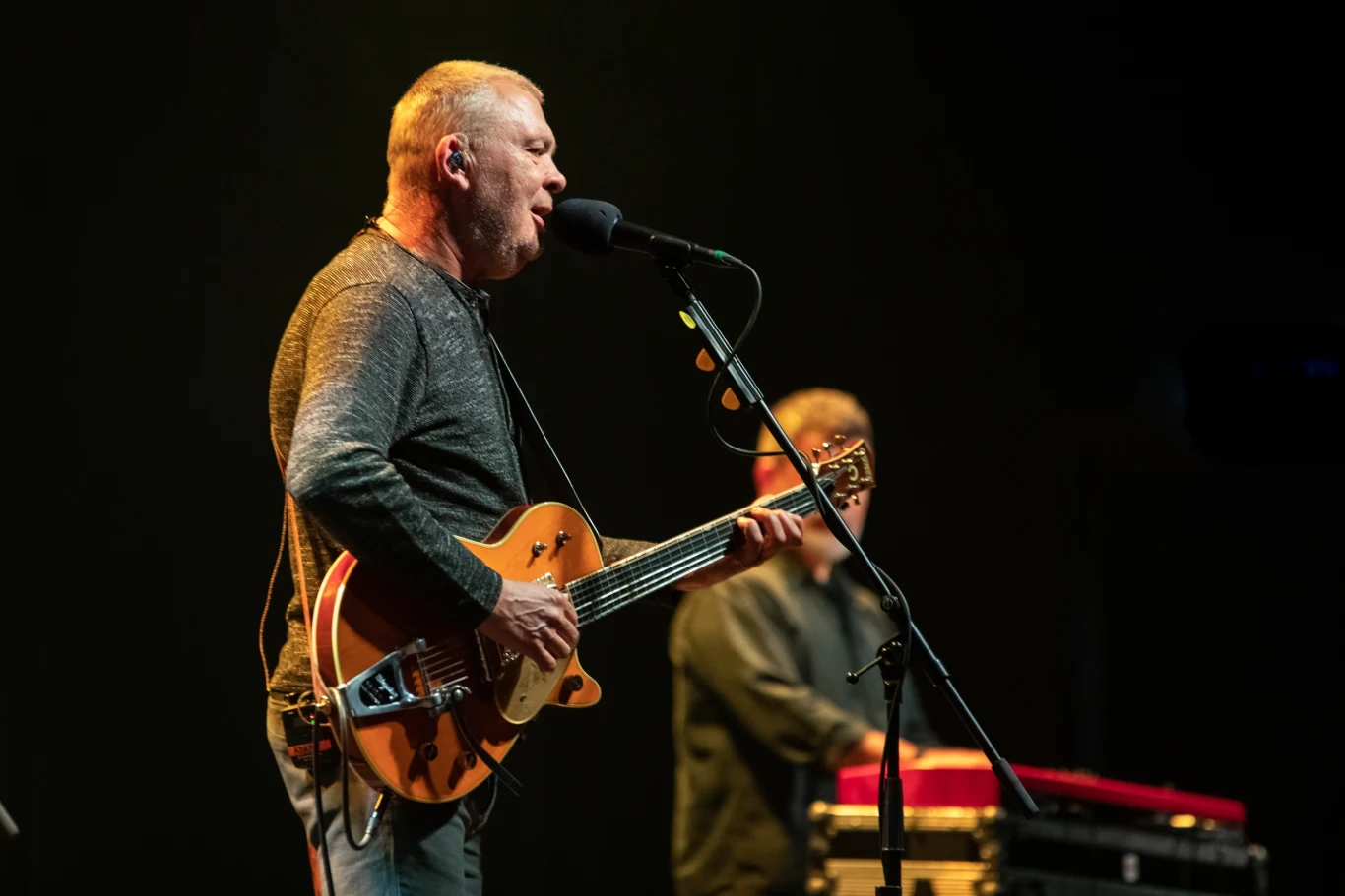 Mężczyzna śpiewający i grający na gitarze elektrycznej na scenie podczas koncertu, w tle drugi mężczyzna przy klawiaturze.