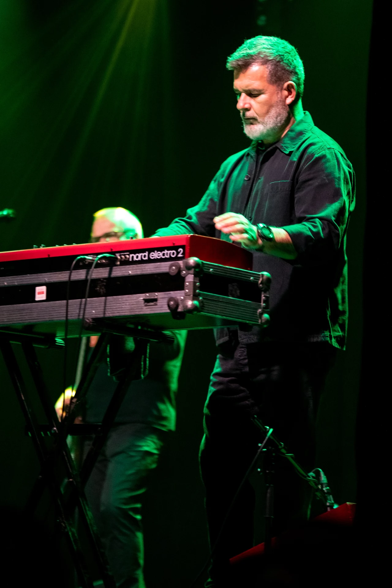 Mężczyzna grający na keyboardzie Nord Electro 2 na scenie, w tle zielone oświetlenie. Koncentracja pianisty widoczna w jego postawie. Muzyka na żywo, koncertowa atmosfera.