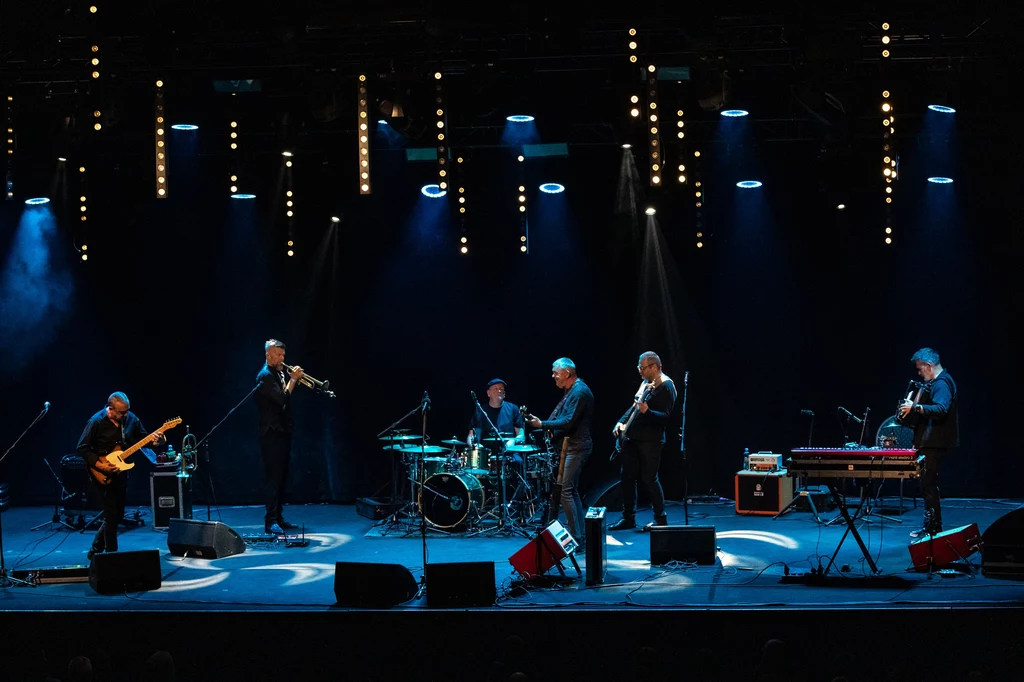 Grupa muzyków grająca koncert na oświetlonej scenie, różne instrumenty takie jak gitara, trąbka i perkusja, wokół ciemne tło i reflektory skierowane na scenę.