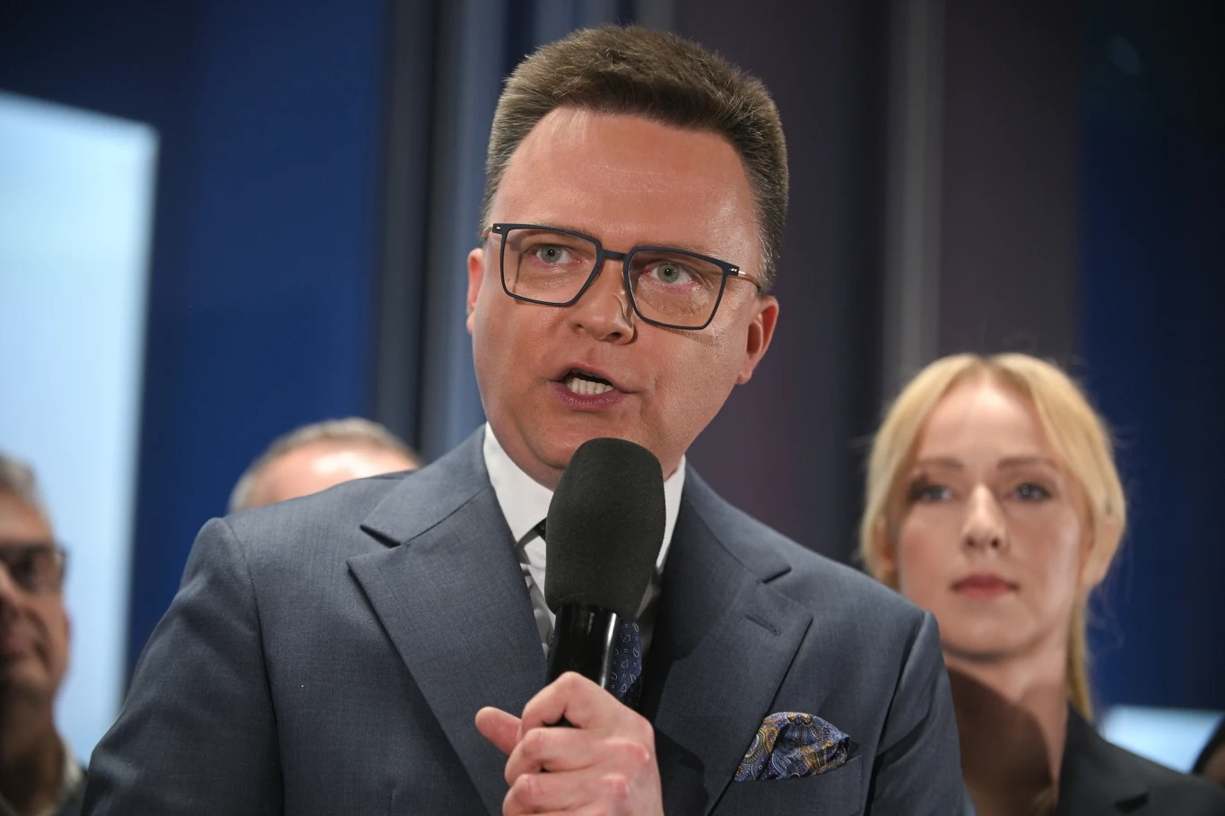 Szymon Hołownia komentuje wyniki sondaży exit poll Mężczyzna w okularach, ubrany w elegancki garnitur, trzyma mikrofon i przemawia, stojąc przed grupą ludzi. Kobieta w blond włosach znajduje się za nim, skupiona na jego słowach. Tło jest zamazane, co podkreśla ich obecność.