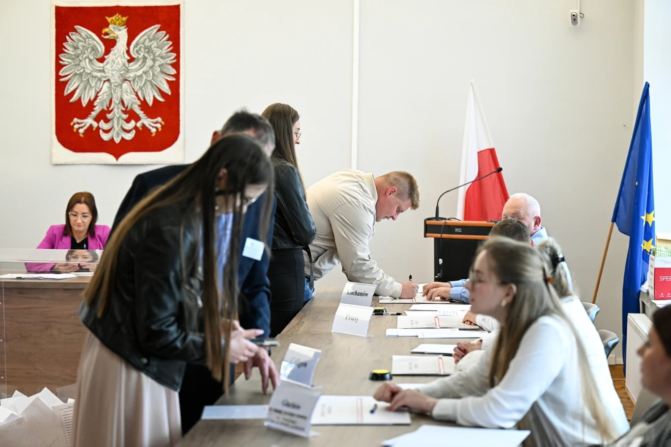 Frekwencja w wyborach prezydenckich według sondażu exit poll Ludzie siedzący przy długim stole w sali konferencyjnej podczas spotkania administracyjnego, w tle widać godło Polski oraz flagę państwową. Uczestnicy są w trakcie zapisywania czegoś na dokumentach.