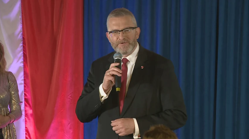 Grzegorz Braun Mężczyzna w garniturze z mikrofonem w ręku podczas wystąpienia publicznego. W tle widać flagę w kolorach białym, czerwonym i niebieskim.