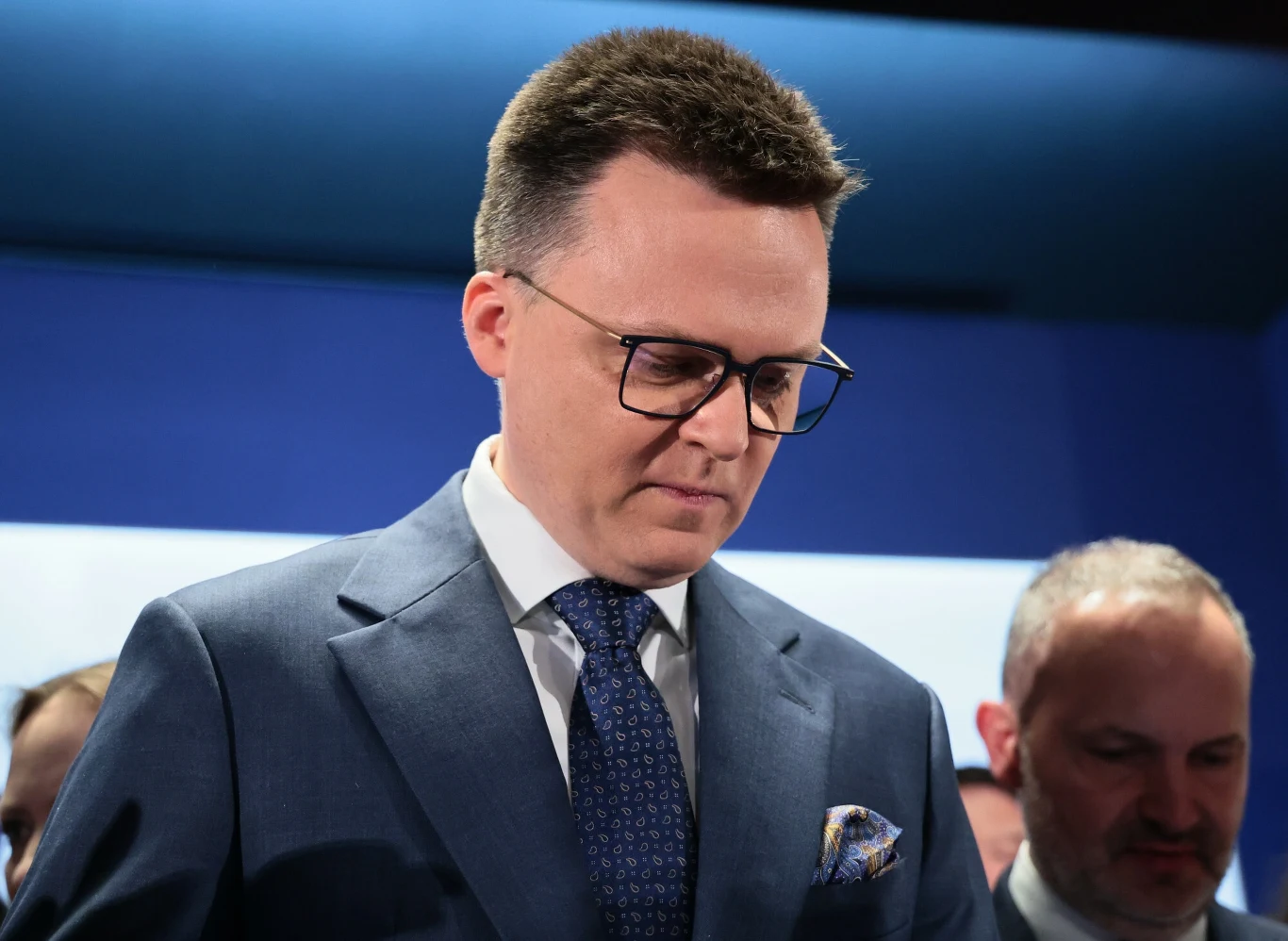 Trzecia Droga z żółtą kartą. Ogromne rozczarowanie w sztabie Hołowni Mężczyzna w okularach i eleganckim garniturze z charakterystyczną poszetką w marynarce. Stoi ze spuszczoną głową, na ciemnym tle.