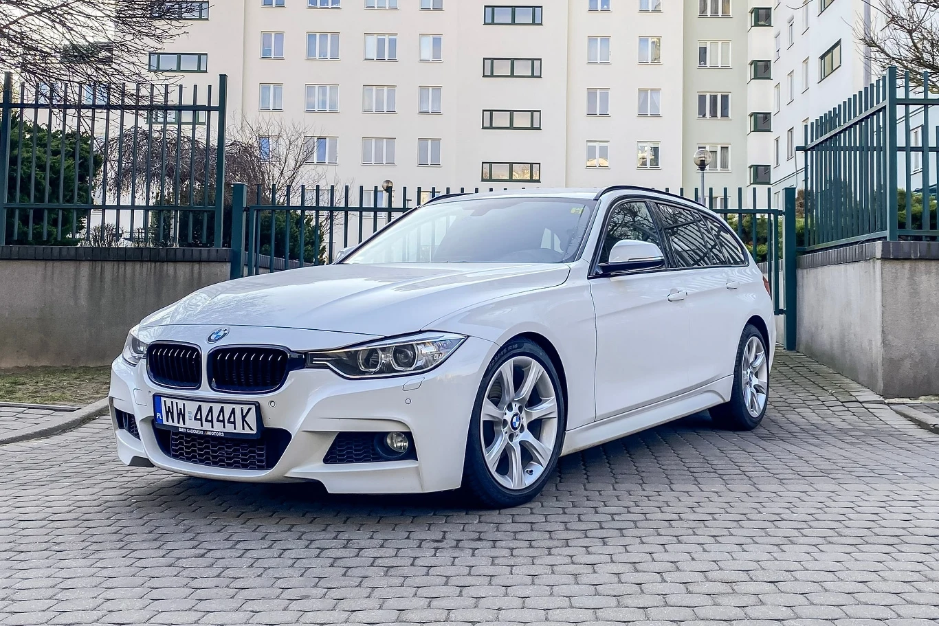 Używane BMW 320d (F31) w wersji Touring prezentuje się atrakcyjnie nawet po latach. Egzemplarz ze zdjęcia ma przebieg ponad 200 tys. km. Został doposażony w fabryczny pakiet M i chiptuning, zwiększający moc do 230 KM. Samochód BMW w kolorze białym zaparkowany na kostce brukowej przed wysokim, nowoczesnym budynkiem.