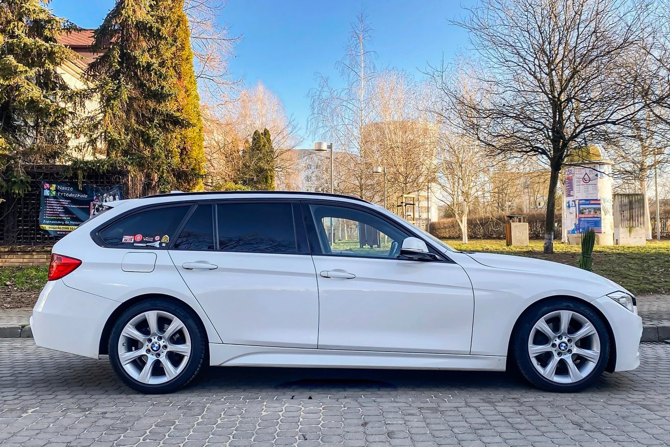 Felgi 18-calowe i opony 225/45 R18 nadają używanemu BMW 320d (F31) sportowego charakteru. Właściciel zrezygnował z run-flatów na rzecz klasycznego ogumienia – to kompromis między komfortem a kosztami. Biały samochód typu kombi zaparkowany na kostce brukowej przy ulicy, w tle widać drzewa i fragmenty budynków. Słoneczny dzień, bezchmurne niebo.