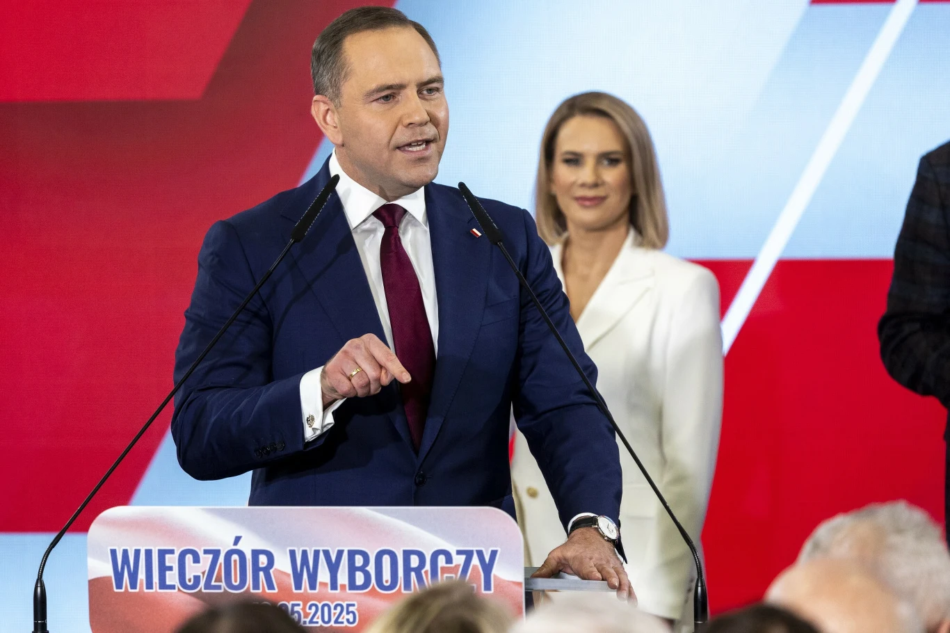 W PiS panuje zadowolenie z wyniku Karola Nawrockiego w I turze wyborów Mężczyzna w garniturze i krawacie przemawia przy mównicy z napisem 'Wieczór Wyborczy 2025'. Kobieta w białej marynarce stoi obok niego na tle w kolorach czerwono-biało-niebieskich. W tle widoczne są zarysy innych osób.