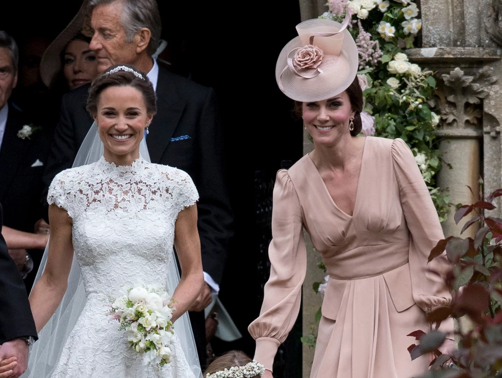 James Middleton mógł liczyć na swoje siostry - Pippę i Kate - gdy sam zmagał się z chorobą Dwie kobiety w eleganckich strojach, jedna w sukni ślubnej z bukietem kwiatów, druga w eleganckim różowym kostiumie z kapeluszem, wychodzą z zabytkowego kościoła ozdobionego kwiatami.