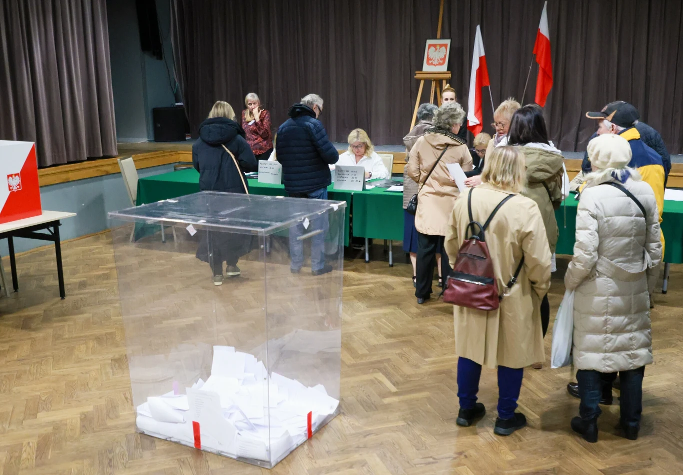 PKW podała dane frekwencyjne Lokal wyborczy z urną wyborczą pełną kart do głosowania na pierwszym planie. W tle widać osoby oddające głosy przy stołach oraz flagi Polski ustawione w pomieszczeniu.