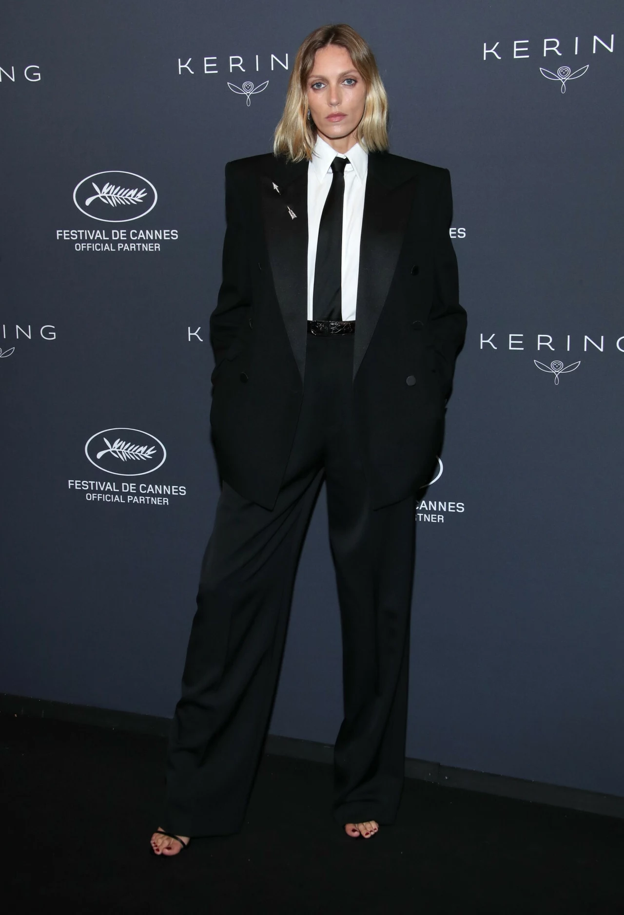 Anja Rubik na festiwalu w Cannes 2025. Modelka pojawiła się na imprezie Kering Women in Motion Osoba ubrana w elegancki czarny garnitur z białą koszulą i krawatem na tle ściany z logo Festiwalu Filmowego w Cannes i Kering.