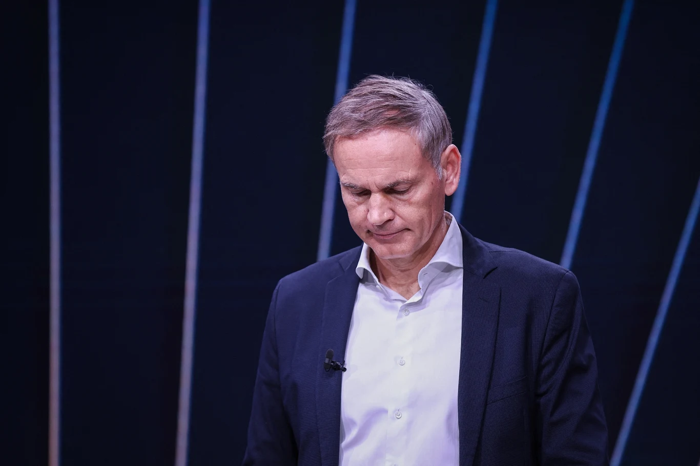 Mężczyzna w garniturze z opuszczoną głową, na ciemnym tle z jasnymi liniami. Na klapie kołnierza ma zamocowany mikrofon. Postawa sugeruje zamyślenie lub skupienie.