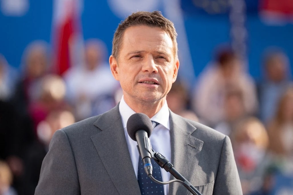 Rafał Trzaskowski Mężczyzna w garniturze przemawia do mikrofonu, na tle rozmytej publiczności i niebieskiego banera. Jego wyraz twarzy jest poważny, a słońce oświetla scenę, tworząc jasną atmosferę.
