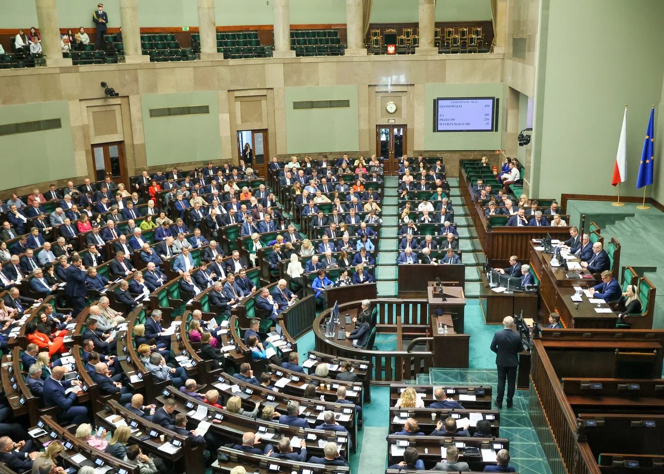 Obrady Sejmu, zdjęcie ilustracyjne Widok na salę obrad zapełnioną osobami siedzącymi na ławkach, skupionymi na przemówieniu trwającym na mównicy. Widoczne flagi Polski i Unii Europejskiej, a nad mównicą zegar i tablica elektroniczna.