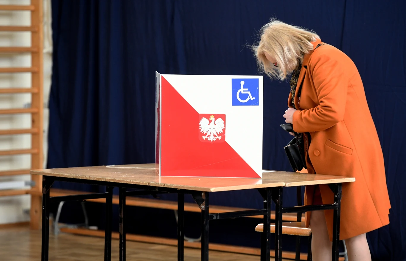 Wybory 2025. Tak głosowano w województwie podlaskim Kobieta w pomarańczowym płaszczu głosuje, pochylona nad urną wyborczą z symbolem orła białego i znakiem dostępności, umieszczoną na drewnianym stole w budynku z ciemnym tłem.