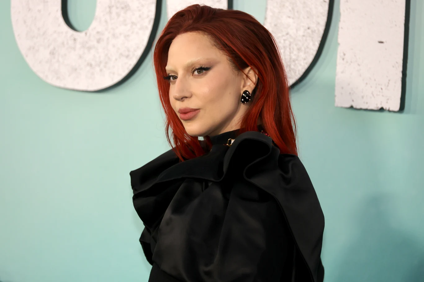 Lady Gaga wystąpi podczas imprezy Netflixa Osoba z długimi, czerwonymi włosami, ubrana w elegancką czarną suknię, pozuje na tle jasnoniebieskiej ściany z dużymi literami.