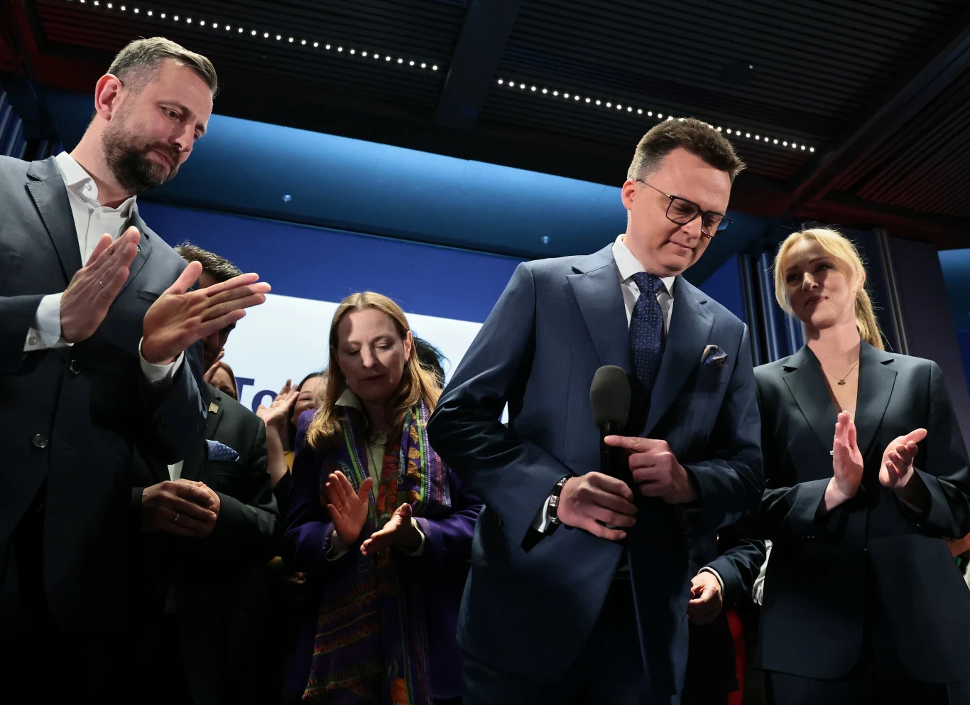 Wieczór wyborczy w sztabie Szymona Hołowni (drugi od prawej). Po lewej Władysław Kosiniak-Kamysz, pierwsza od prawej - Urszula Brzezińska-Hołownia Grupa ludzi w eleganckich strojach, skupiona na osobie trzymającej mikrofon. W tle widoczny jest wystrój sceniczny. Osoby na zdjęciu wyglądają na zaangażowane w wydarzenie, być może konferencję prasową lub przemówienie.