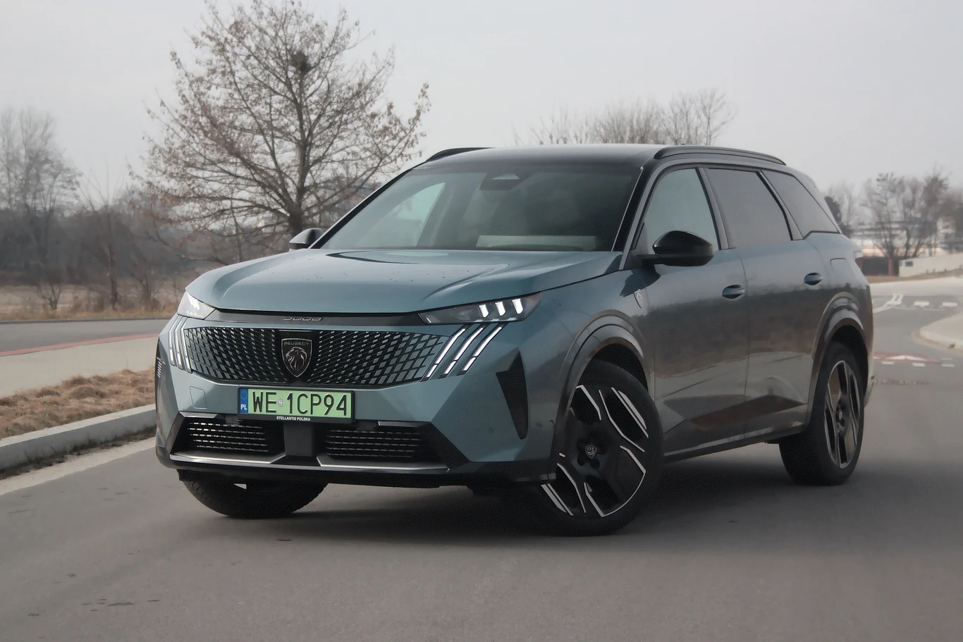 Peugeot E-5008 to siedmiomiejscowy samochód o napędzie elektrycznym Samochód SUV o nowoczesnym wyglądzie, zaparkowany na bocznej drodze. Ma charakterystyczne przednie światła LED i polską tablicę rejestracyjną. W tle widać bezlistne drzewa i jasne niebo.