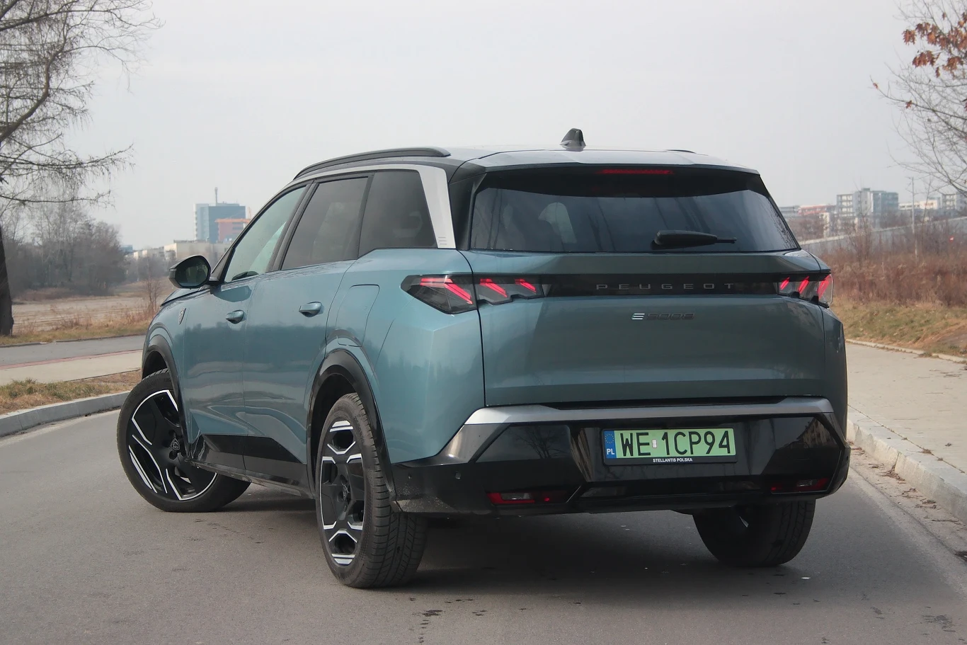 Tył Peugeota e-5008 nie jest tak efektowny jak przód, ale i tu styliści wycisnęli, ile się dało Zdjęcie przedstawia nowoczesny SUV z zielonymi tablicami rejestracyjnymi, stojący na asfaltowej drodze na tle miejskiego krajobrazu z drzewami i budynkami w oddali.