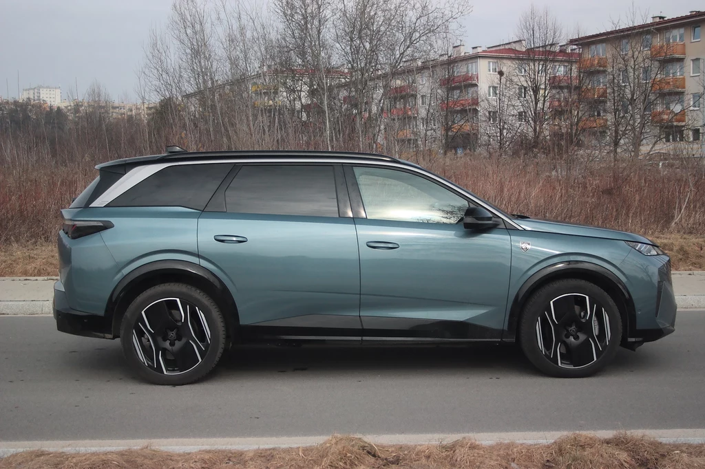 Stylistyka boku Peugeota e-5008 jest stonowana Samochód SUV zaparkowany na ulicy, w tle widać osiedle z wielopiętrowymi blokami mieszkalnymi i drzewa bez liści. Auto ma niebieski lakier i nowoczesny design, wyróżnia się masywnymi kołami i aerodynamiczną sylwetką.