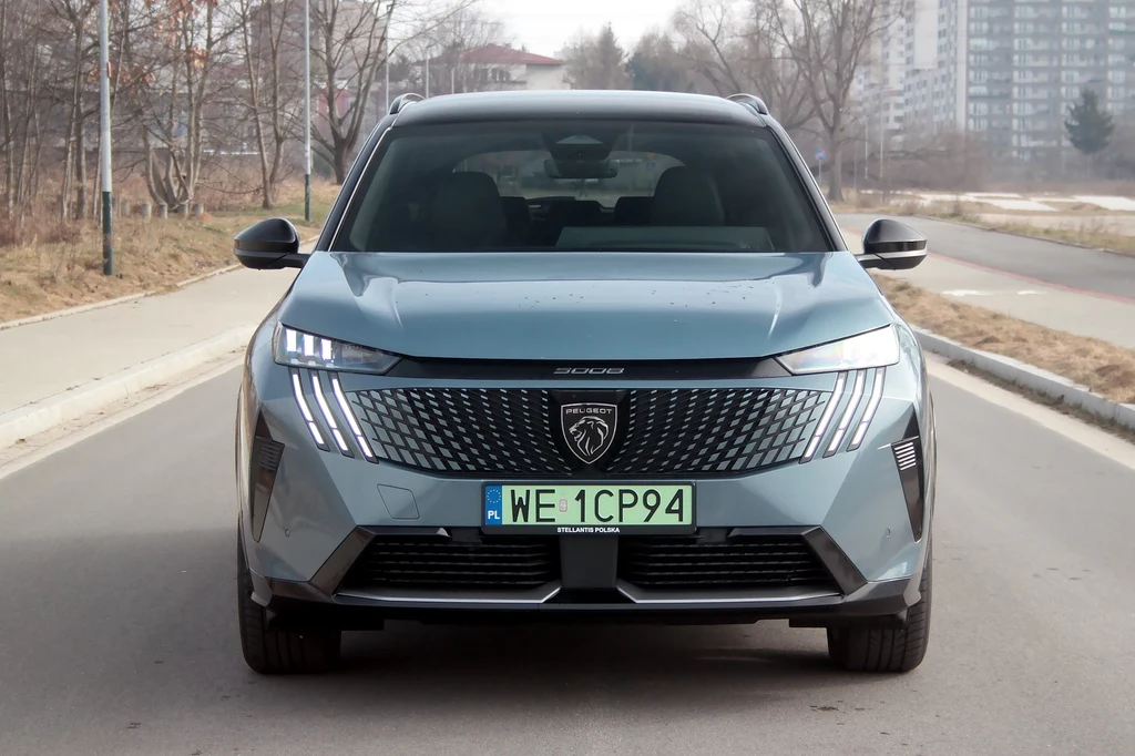 Najefektowniej Peugeot e-5008 wygląda z przodu Nowoczesny samochód osobowy marki Peugeot stoi na pustej drodze miejskiej, z wyraźnym widokiem na tablicę rejestracyjną. W tle widać budynki i zimowy krajobraz.