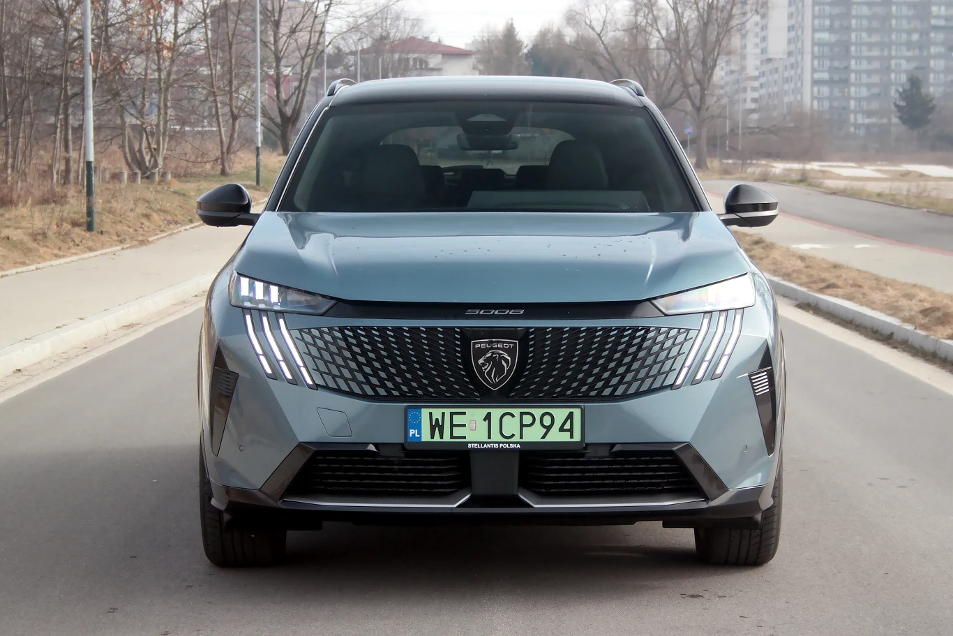 Najefektowniej Peugeot e-5008 wygląda z przodu Nowoczesny samochód osobowy marki Peugeot stoi na pustej drodze miejskiej, z wyraźnym widokiem na tablicę rejestracyjną. W tle widać budynki i zimowy krajobraz.