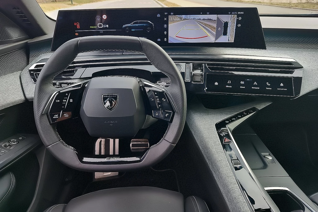 Cały kokpit Peugeota e-5008 wygląda futurystycznie Wnętrze luksusowego samochodu z nowoczesnym panelem sterowania. Widoczna kierownica ze specjalistycznymi przyciskami oraz cyfrowy wyświetlacz pokazujący informacje o pojeździe. Deska rozdzielcza ma minimalistyczny, elegancki design.