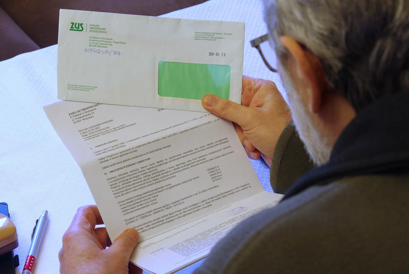 Kończy się świadczenie rehabilitacyjne - co dalej? Mężczyzna trzyma w rękach dokument oraz kopertę z logo ZUS. Dokument znajduje się na pierwszym planie i zawiera tekst. Na stole leżą również inne przedmioty, takie jak długopisy.