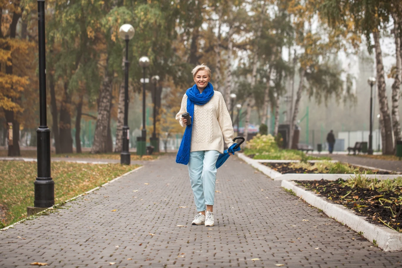 Kobieta spacerująca po parku jesienią, ubrana w beżowy sweter, niebieski szalik i spodnie. Trzyma kubek z kawą i kurtkę. W tle widać drzewa z żółtymi liśćmi i latarnie ustawione wzdłuż alejki.