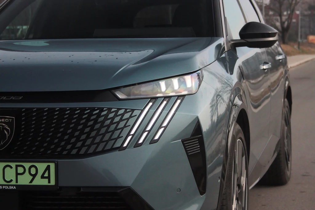 Peugeot e-5008 Nowoczesny samochód osobowy w kolorze niebieskim, widok na przód pojazdu z wyeksponowanymi przednimi światłami LED i charakterystycznym grillem. Widoczna jest także tablica rejestracyjna z zieloną ramką. Auto stoi na drodze.