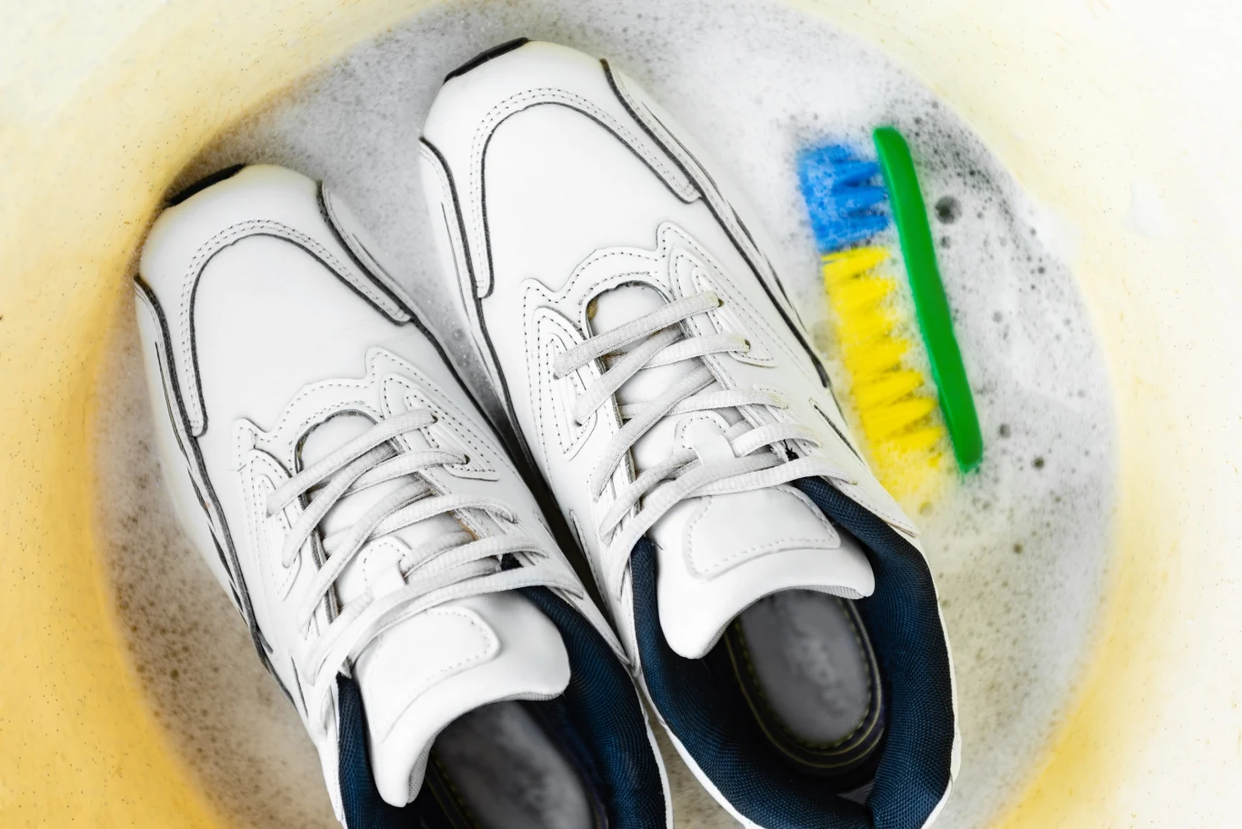 Białe buty sportowe zanurzone w wodzie z pianą, obok znajduje się plastikowa szczotka do czyszczenia o zielonej rączce i kolorowym włosiu.