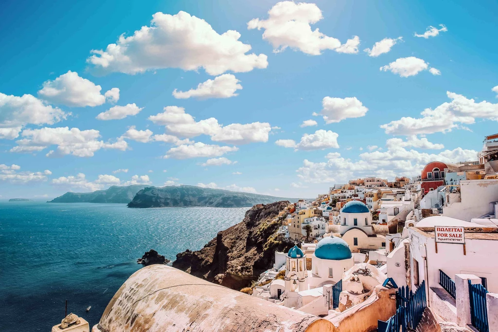 All Inclusive i Last Minute, przepis na udany wyjazd Białe budynki z niebieskimi kopułami położone na klifie nad lazurowym morzem, charakterystyczna architektura Santorini przy idealnie błękitnym niebie.