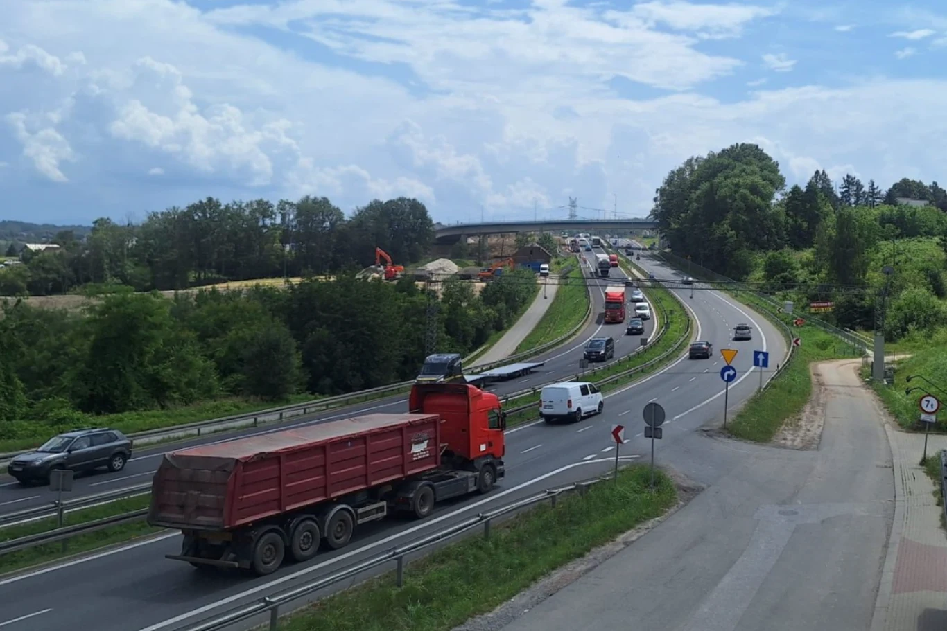 Dziś w nocy rozpocznie się kolejny etap prac, który potrwa przez 4 tygodnie. Autostrada z licznymi pojazdami w ruchu, w tym ciężarówka oraz samochody osobowe, otoczona zielenią i drzewami, z mostem w oddali i pod chmurnym niebem.