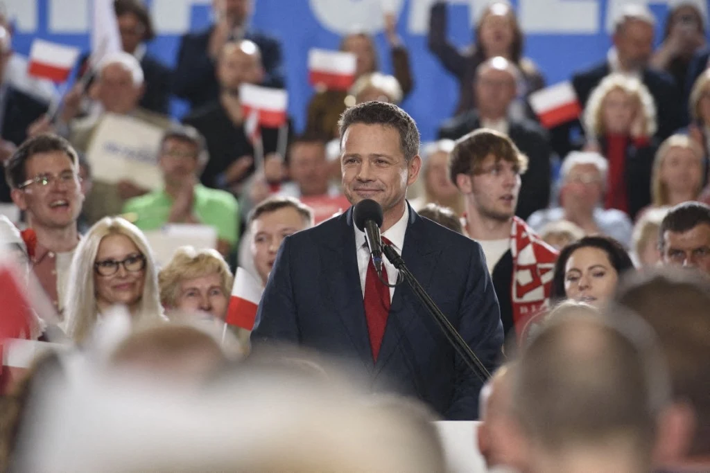 Polityk w garniturze przemawia do tłumu, trzymając mikrofon. W tle liczna publiczność z flagami Polski. Atmosfera wydarzenia jest ożywiona i pełna entuzjazmu.