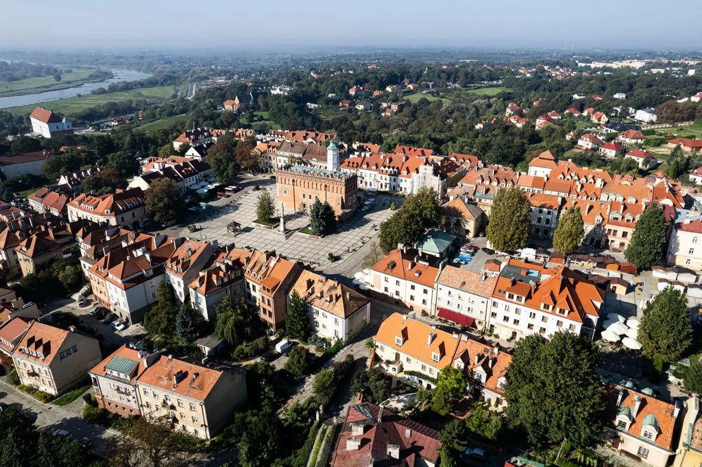 Sandomierz zachwyca architekturą Widok z lotu ptaka na zabytkowe centrum miasta, z pięknym rynkiem i otaczającymi go kamienicami z czerwonymi dachami. W tle rzeka i rozległe tereny zielone podkreślają malowniczy charakter miejsca.