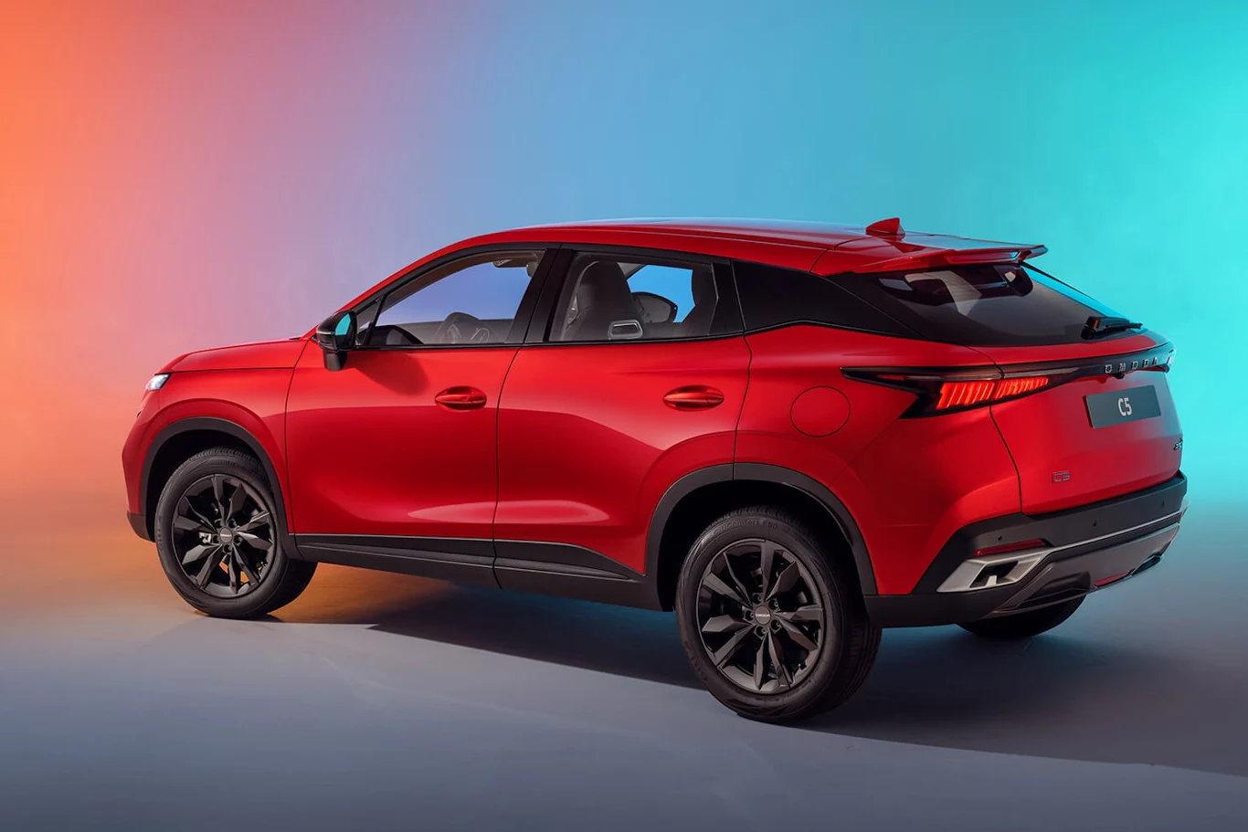 Omoda 5 po liftingu Czerwony SUV na tle gradientowego światła z przejściem od pomarańczowego do niebieskiego. Modernistyczny design z wyraźnymi, ostrymi liniami oraz matowymi, czarnymi felgami podkreśla nowoczesność pojazdu.