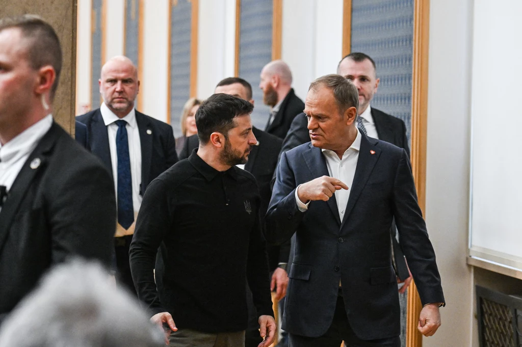 Prezydent Ukrainy Wołodymyr Zełenski i premier Polski Donald Tusk Dwóch mężczyzn ubranych w eleganckie stroje rozmawia w korytarzu otoczonym przez innych ludzi. Jeden z mężczyzn gestykuluje ręką, podkreślając wagę rozmowy.