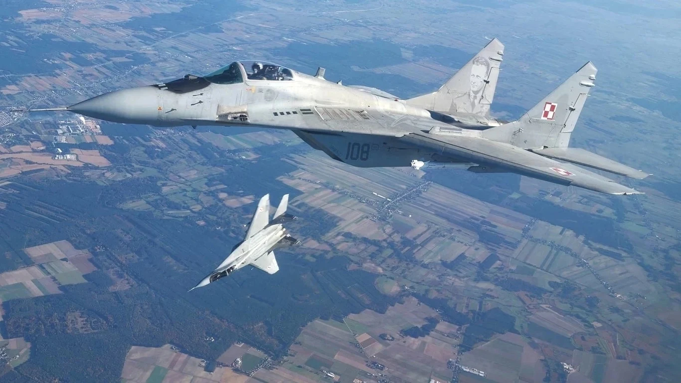 Ukraińcy zaatakowali Rosjan polskim myśliwcem i bronią z USA Dwa wojskowe myśliwce Mig-29 w locie nad krajobrazem wiejskim, widoczna z góry różnorodność pól i lasów. Na jednym z samolotów widnieje polska flaga.