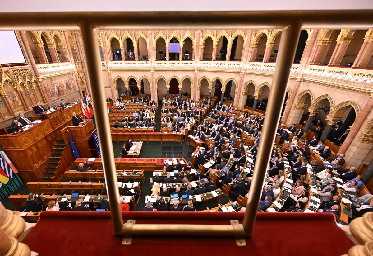 Sala obrad węgierskiego parlamentu Wnętrze sali obrad parlamentu, widok z galerii nad salą. Dookoła ozdobne łuki i kolumny, w centrum posłowie siedzący przy stołach z dokumentami. Na lewo podium dla prowadzącego, obok flagi państwowe. Atmosfera formalna i uroczysta.