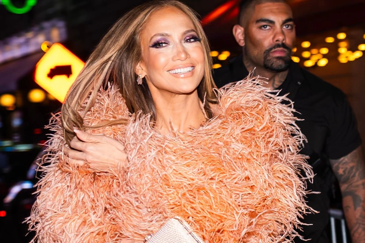 Jennifer Lopez zachwyciła, pojawiając się na ulicach Nowego Jorku Elegancko ubrana kobieta w pomarańczowym futrze z piórami, uśmiechnięta, z długimi rozpuszczonymi włosami. W tle widać stojącego ochroniarza. Neonowe światła w tle dodają blasku scenie.