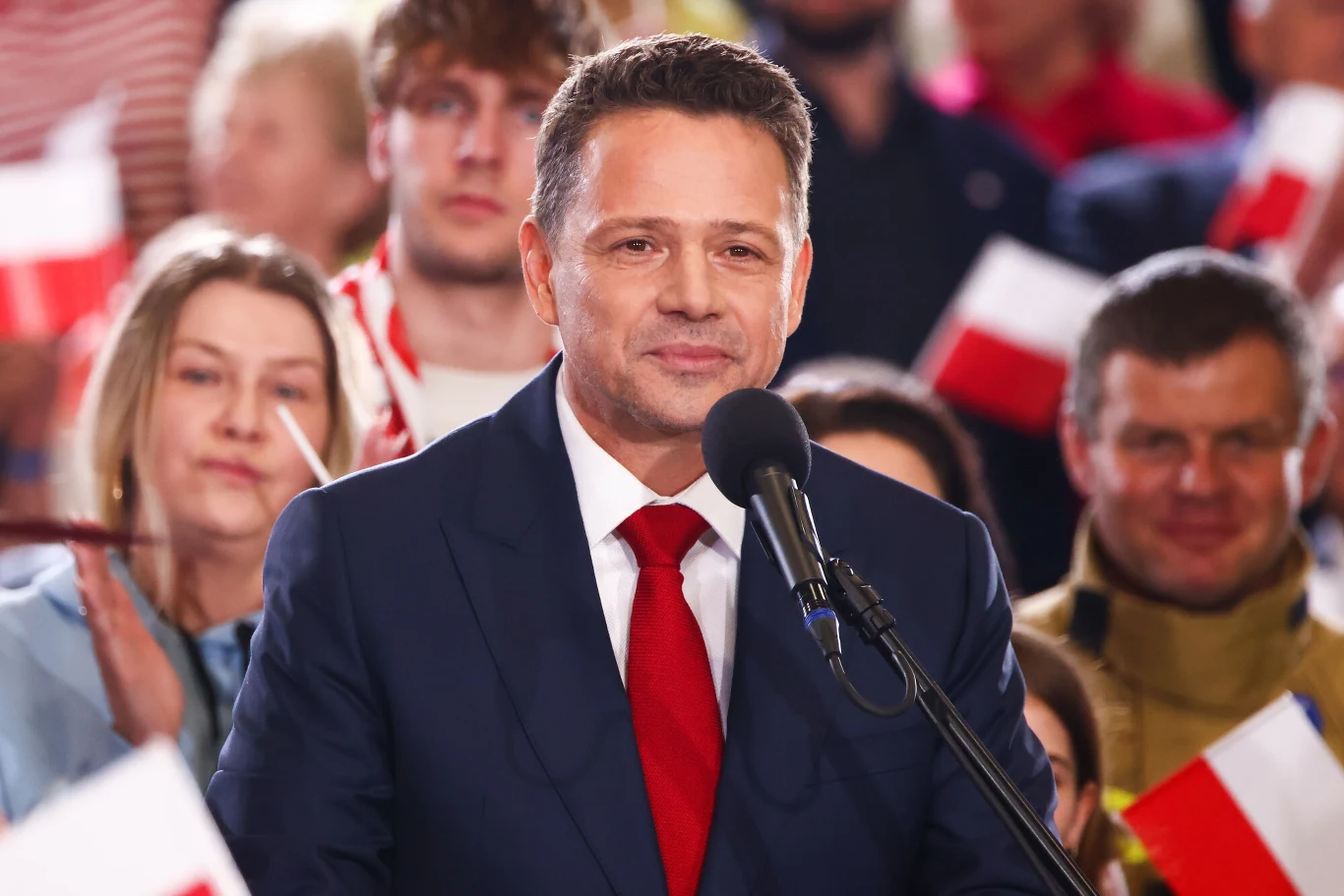 Mężczyzna w garniturze z czerwonym krawatem przemawia do zgromadzenia, trzymając mikrofon. W tle widoczny tłum ludzi z biało-czerwonymi flagami.