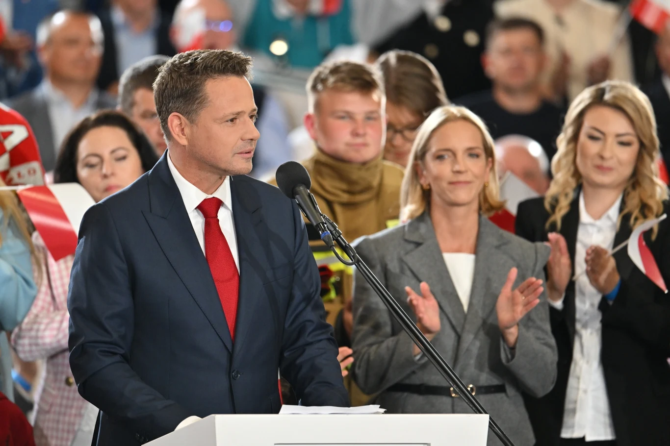 Niemcy piszą o wyborach prezydenckich w Polsce. Na zdj. Rafał Trzaskowski Osoba w garniturze przemawia przy mikrofonie na wydarzeniu publicznym, w otoczeniu klaskających ludzi. W tle widoczne są flagi i skupiona publiczność.