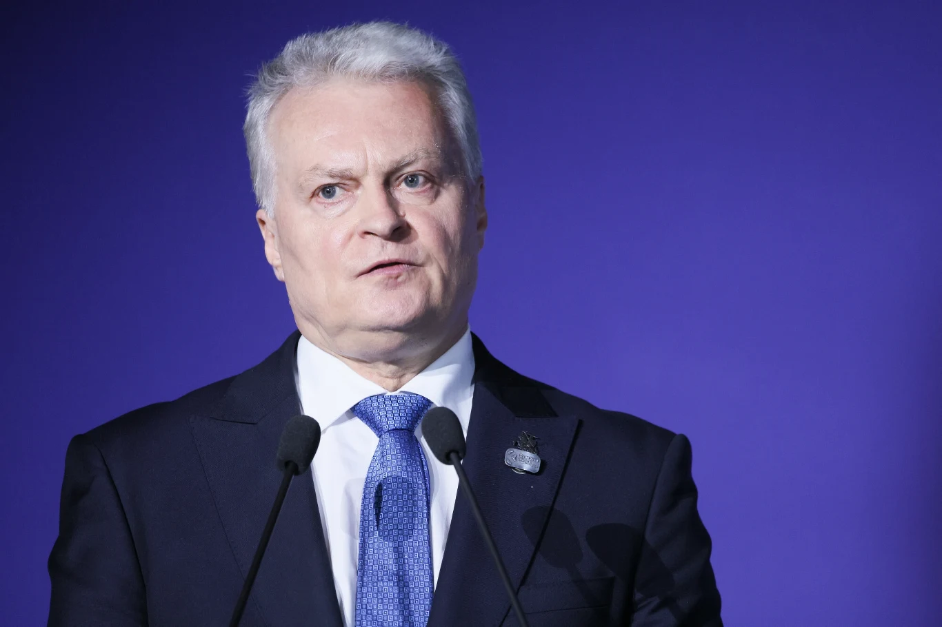 Prezydent Gitanas Nauseda komentuje wybory prezydenckie w Polsce. Mężczyzna ubrany w formalny garnitur i niebieski krawat z przypinką na klapie, stoi przed dwoma mikrofonami na tle niebieskiej ściany.